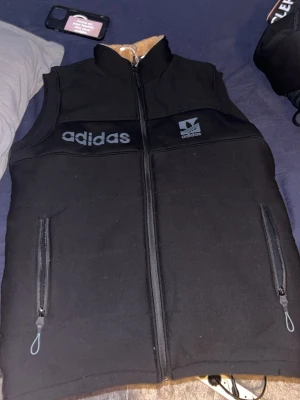 Svart väst från Adidas - Svart väst från Adidas med dragkedja framtill och två fickor med dragkedja. Adidas-logga och text på både fram- och baksida. Västen har hög krage och är fodrad på insidan för extra värme.