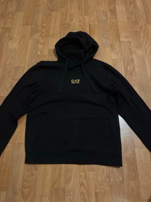 Svart hoodie från EA7 med gul logga - Svart hoodie från EA7 med gul logotyp på bröstet och gul detalj på ryggen. Klassisk modell med huva och dragsnören samt en stor magficka framtill. Perfekt för en stilren och sportig look.