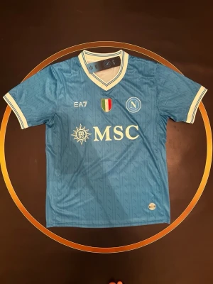 Napoli matchtröja blå EA7 - Snygg ljusblå Napoli matchtröja från EA7 med vita detaljer och klubbmärke på bröstet. Tröjan har V-ringning, broderad Scudetto och MSC-logga framtill. Perfekt för fotbollsfans som vill sticka ut på läktaren.