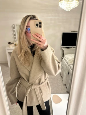 Beige omlottkappa med bälte - Stilren beige kappa i omlottmodell med bälte i midjan. Kappan har en avslappnad passform och långa ärmar. Super skön och fin till båda vardaglig eller speciella tillfällen.