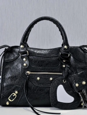 Balenciaga city bag - Cool svart handväska i skinn med guldfärgade detaljer, dragkedja framtill och dekorativa nitar. Väskan har dubbla handtag och en avtagbar spegel med hjärtformad detalj. Perfekt för dig som gillar edgy accessoarer.