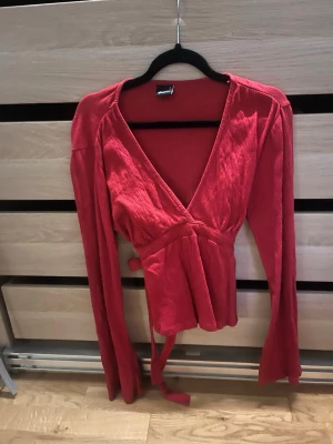 Röd omlottopp med knytband - Trendig röd omlottopp med långa ärmar och knytband i midjan. Toppen har en djup v-ringning och är tillverkad i ett mjukt, lätt material som ger en snygg passform. Perfekt för dig som vill ha en stilren men ändå iögonfallande look.