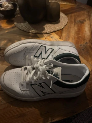 Vita New Balance sneakers med gröna detaljer - Snygga vita sneakers från New Balance med gröna detaljer och klassisk N-logga på sidan. Skorna har snörning, perforerad tå och en låg siluett. Perfekta för dig som gillar stilrena och sportiga sneakers.