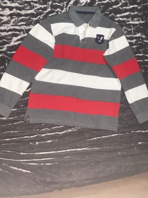 Randig långärmad rugbytröja - Snygg långärmad rugbytröja med breda ränder i grått, vitt och rött. Tröjan har krage, knappar vid halsen och ett broderat märke med örn på bröstet. Perfekt för en sportig och avslappnad stil.