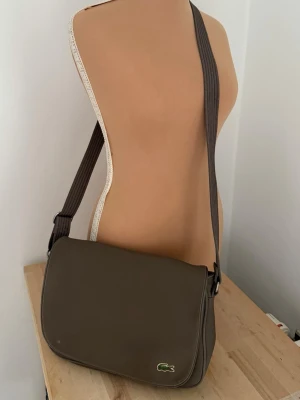 Brun axelväska från Lacoste - Snygg brun axelväska från Lacoste med klassisk krokodillogga framtill. Väskan har ett justerbart axelband och lock med magnetknappar. Insidan av locket har ett grönt och vitt randigt mönster. Perfekt för att bära dina viktigaste grejer.