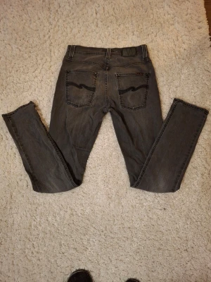 Grå jeans från Nudie Jeans - Snygga grå jeans från Nudie Jeans med klassisk femficksdesign och svarta sömmar på bakfickorna. Jeansen har raka ben och normal passform, perfekta till en avslappnad stil. Tillverkade i slitstarkt bomullsmaterial.