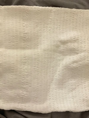 Vit smockad bandeau tubtopp - Vit bandeau tubtopp med smockad struktur och stretchigt material. Toppen är ärmlös och har en enkel, stilren design som passar perfekt till sommaren. Materialet känns mjukt och följsamt mot huden.