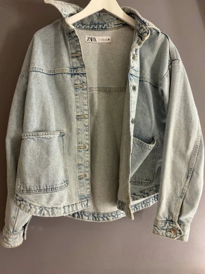 Ljusblå jeansjacka från Zara - Säljer en ljusblå jeansjacka från Zara med oversized passform. Jackan har stora fickor framtill, klassisk krage och knappar längs hela framsidan. Perfekt att slänga över en hoodie eller t-shirt för en chill look.