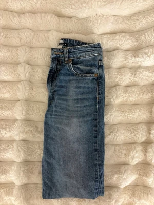 Blå raka jeans i klassisk modell - Säljer ett par blå jeans med rak passform och klassisk femficksdesign. Jeansen har normalhög midja, bälteshällor och är tillverkade i denim. Perfekta för en avslappnad och tidlös stil.