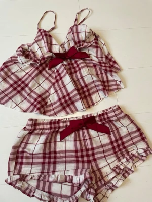 Rutigt pyjamasset med rosetter - Supersött pyjamas-set med linne och shorts i röd- och vitrutigt mönster. Båda delarna har dekorativa rosetter framtill och volangdetaljer längs kanterna. Perfekt för mysiga hemmakvällar! Materialet känns mjukt och luftigt.