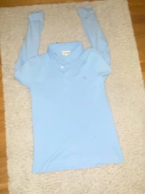 Ljusblå långärmad polotopp Lacoste - Snygg ljusblå långärmad polotopp från Lacoste med klassisk krage och knappar framtill. Ikoniska krokodilloggan broderad på bröstet. Perfekt till jeans eller kjol för en clean och sportig look.