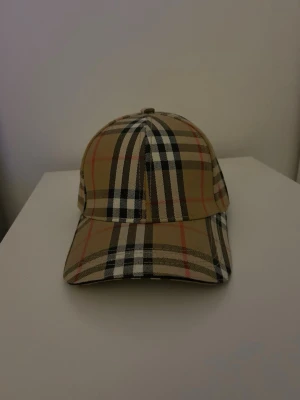 Beige keps med rutigt mönster Burberry - Säljer en klassisk beige keps från Burberry med det ikoniska rutiga mönstret i svart, vitt och rött. Kepsen har böjd skärm och justerbart spänne bak i metall med Burberry-logga. Tillverkad i bomullstyg och perfekt för dig som gillar stilrena accessoarer. Helt ny aldrig använd!