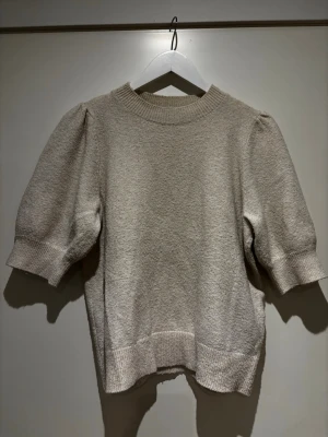 Beige stickad topp med puffärm  - Trendig beige topp med puffärmar och ribbade muddar. Kortare ärm och rund halsringning ger en modern look. Materialet har en lätt struktur som gör tröjan extra intressant.