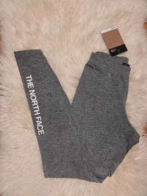 Grå leggings från The North Face - Snygga grå leggings från The North Face med vit logga längs ena benet. De är tillverkade i ett stretchigt och mjukt material som passar perfekt för träning eller chill. Hög midja och smal passform ger en modern look.