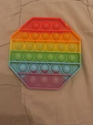 Regnbågsfärgad pop it-leksak - Säljer en åttakantig pop it-leksak i regnbågens färger. Den är gjord av mjuk silikon och har flera rader med bubblor att trycka på. Perfekt för att sysselsätta händerna eller bara för att slappna av.