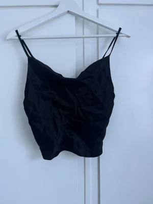 Svart satinlinnetopp med smala band - Snygg svart linnetopp i satin med dubbla smala axelband. Toppen har en lätt glansig finish och en enkel, croppad passform som passar perfekt till både jeans och kjol. Perfekt för dig som gillar minimalistisk stil.