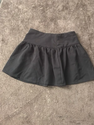 Svart kjol med låg midja - Snygg svart kjol med låg midja och utsvängd kjoldel. Skortsen har en bred linning och är tillverkade i ett mjukt, följsamt material som ger en bekväm passform. Perfekta för dig som vill ha kjol-looken men ändå känna dig bekväm.