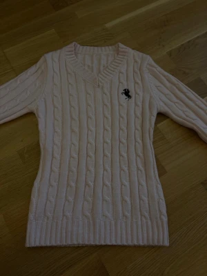 Ljusrosa kabelstickad tröja Ferrari - Ljusrosa kabelstickad tröja från Ferrari med V-ringning och svart broderad logotyp på bröstet. Tröjan har långa ärmar och ribbade muddar vid ärmslut och nederkant. Perfekt för dig som gillar klassisk stil med en sportig touch.