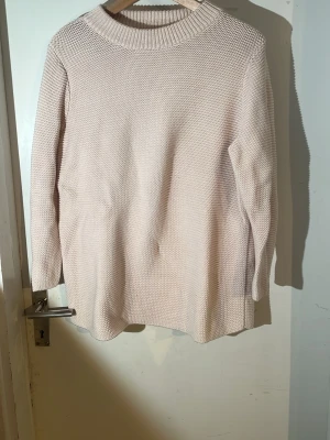 Ljusrosa/vit stickad tröja - En oversized, ljusbeige stickad tröja med grov struktur och rund halsringning. Tröjan har långa ärmar och bred ribbad kant vid halsen. Perfekt för lager på lager och enkel att matcha med olika stilar. Materialet ser ut att vara mjukt och bekvämt, troligen bomull eller bomullsblandning.