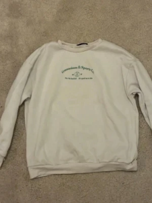 Vit sweatshirt med grön text - Vit sweatshirt med rund hals och tryck i grönt framtill där det står 'Gymnasium & Sports Co.'. Tröjan har en avslappnad passform  och ribbade muddar vid ärmar och nederkant. Perfekt för en chill och sportig look.