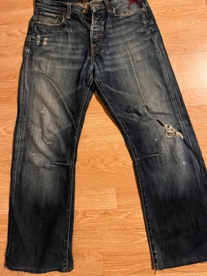 Äkta Ed Hardy Jeans - Unika blå jeans med slitningar och broderade döskallar på bakfickorna. Raka ben och klassisk femficksmodell. Snygg tvättad look och detaljer som ger extra edge. Perfekta för dig som vill sticka ut med coola detaljer.