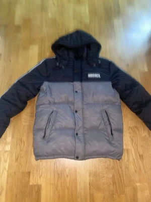 Grå och svart pufferjacka Hoodrich - Säljer en snygg pufferjacka från Hoodrich i grått och svart med huva och dragkedja framtill. Jackan har två sidofickor med dragkedja och logga på bröstet. Perfekt för kalla dagar och riktigt street.