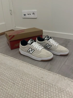 Beige New Balance sneakers med vit sula - Snygga beige New Balance sneakers med vit sula och bruna detaljer. Skorna har klassisk snörning, svart innerfoder och den ikoniska N-loggan på sidan. Ovandelen är i mocka och mesh för en sportig och fräsch look.