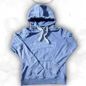 Blå hoodie från Superdry - Snygg ljusblå hoodie från Superdry med stor magficka och vit dragsko i huvan. Tryckt Superdry-logga framtill och ribbade muddar vid ärmar och nederkant. Perfekt för en avslappnad och trendig look. Passar någon 165-175