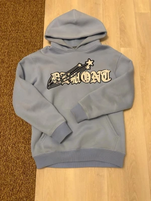 Ljusblå hoodie från Demont XS - Snygg ljusblå hoodie från Demont i storlek XS. Hoodien har en stor huva och ett broderat motiv med texten 'Demont' och en stjärnshooting detalj på bröstet. 