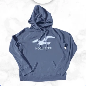 Blå hoodie från Hollister - Mörkblå hoodie från Hollister med stor logga och fågelmotiv på bröstet. Klassisk känguruficka framtill och justerbar dragsko i huvan. Perfekt för en avslappnad och cool stil. Passar 165-175