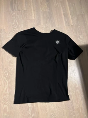 Svart Stone Island t-shirt - Snygg svart t-shirt från Stone Island med klassisk logopatch på bröstet. T-shirten har rund hals och korta ärmar, tillverkad i mjuk bomull för skön känsla. Perfekt för dig som gillar stilrena och tidlösa plagg.
