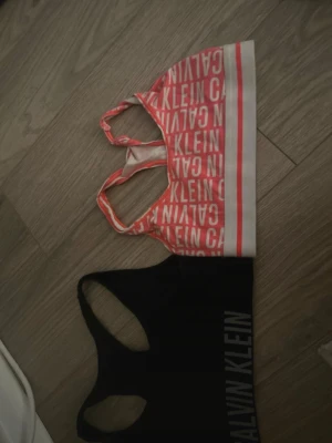 Två Calvin Klein sport-bhar - Säljer två sportiga bhar från Calvin Klein. Den ena är vit och korallröd med logotypmönster och bred resår, den andra är helsvart med Calvin Klein-tryck i vitt på sidan. Båda har brottarrygg och är tillverkade i mjukt stretchmaterial.