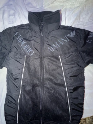 Svart vindjacka från Emporio Armani L/M - Svart vindjacka från Emporio Armani med hög krage och dragkedja framtill. Jackan har stora logotyper och emblem både fram och bak samt vita detaljer längs sidorna. Tillverkad i syntetmaterial med sportig känsla och meshpaneler för extra ventilation.
