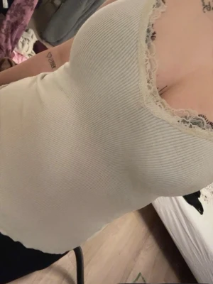 Vit ribbad linnetopp med spetskant - Snygg vit ribbad linnetopp med spetskant runt urringningen. Toppen är ärmlös och har en figurnära passform som framhäver siluetten. Perfekt att styla med jeans eller kjol för en trendig look.