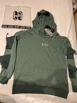 Grön hoodie från HTGY med detaljer - Snygg grön hoodie från HTGY med coola detaljer som snörning och fickor på ärmarna. Tröjan har huva med spännen och ribbade muddar. Materialet är mjukt och känns skönt mot huden, perfekt för dig som gillar streetwear och vill sticka ut.