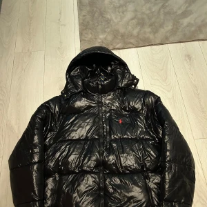 Svart pufferjacka Polo Ralph Lauren M - Svart pufferjacka från Polo Ralph Lauren med glansig finish och röd logga på bröstet. Jackan har huva, dragkedja framtill och flera fickor. Materialet är en blandning av bomull och polyamid, perfekt för kalla dagar. Använd 3-4 ggr 