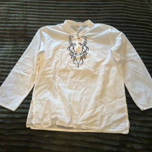 Vit skjorta med broderi - Vit skjorta med svarta broderade detaljer framtill och två träknappar vid halsen. Finns ingen lapp men gissar på storlek M