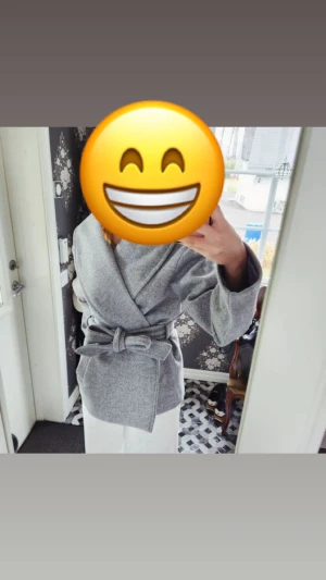 Grå omlott kappa med bälte - Snygg grå kappa i omlottmodell med bälte i midjan och bred krage. Jackan har långa ärmar och två stora fickor framtill. Perfekt för dig som gillar stilrena och enkla plagg med en chill vibe. Materialet känns mjukt och har en ull-liknande look.