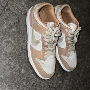 Nike Dunk sneakers i beige och vitt - Nike Dunk sneakers i en snygg kombination av beige mocka och vitt skinn. Låg modell med rund tå, platt sula och klassisk snörning. Swoosh-loggan i ljusgrått på sidan och mjukt foder i beige. Perfekt för dig som gillar stilrena och neutrala färger. Kan tillägga att priset kan diskuteras vid en snabb affär.