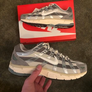 Nike P-6000 grå sneakers strl 43 - Nike P-6000 sneakers i grå och silver med mesh och syntetpaneler, vita detaljer och snörning. Snygg retrostil med rund tå och platt sula, perfekt för dig som gillar sportig vibe och streetwear. Sulan har svarta och ljusblå inslag för extra kontrast.