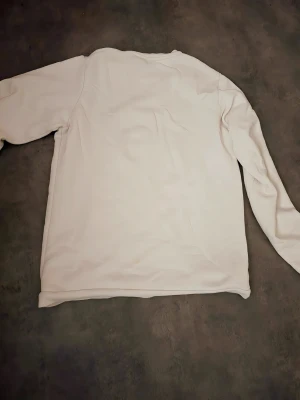 Vit sweatshirt från Lacoste med tryck - Vit sweatshirt från Lacoste med klassiskt grönt krokodiltryck och logga på bröstet. Tröjan har rund hals, långärmade ärmar och är gjord i mjuk bomull.  Storlek är Xs men passar som S Perfekt för dig som gillar stilrena och sportiga plagg med tydlig branding.