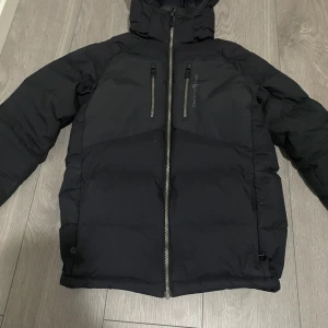 Svart dunjacka Sail Racing 160 - Svart dunjacka från Sail Racing, modell Sail Antarctica Expedition. Jackan har huva, dragkedja framtill och patch på ärmen. Fylld med dun och har en clean, puffad look. Perfekt för kalla dagar och har snygga detaljer som logga och zip på ärmen.