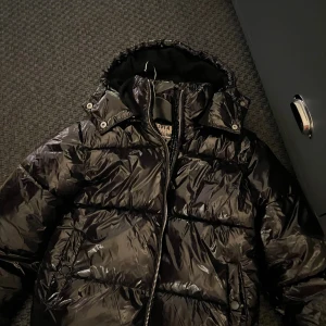Svart glansig pufferjacka med huva - Säljer en svart glansig jacka🥰andvänd förra vintern några få gånger,säljer för jag har andra jackor🥰 har inga tecken på andvändning eller skador.pris kan diskuteras