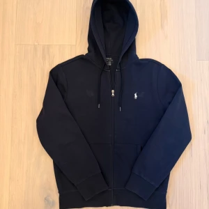 Svart hoodie från Polo Ralph Lauren - Svart hoodie från Polo Ralph Lauren med dragkedja och huva. Tröjan har snörning vid halsen, klassisk logga broderad på bröstet och två fickor framtill. Tillverkad i mjuk bomull, perfekt för chill dagar. Snygg och enkel design som funkar året runt. Den har 2 små fläckar 