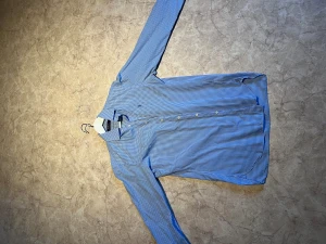 Blårutig skjorta från Polo Ralph Lauren - Snygg blårutig skjorta från Polo Ralph Lauren med klassisk krage och knappar framtill. Skjortan har lång ärm och det ikoniska broderade Polo-logot på bröstet. Tillverkad i mjuk bomull som känns skön mot huden. Perfekt för dig som gillar stilren och tidlös design. Passar L/M