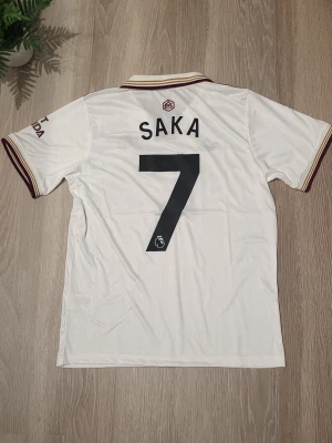 Arsenal Saka trdje kit fotbollströja - Säljer en officiell Arsenal fotbollströja från Adidas med Saka och nummer 7 på ryggen. Tröjan är vit med vinröda detaljer, klassisk krage och korta ärmar. Materialet är lätt och andas, perfekt för match eller träning. Tryck med Emirates Fly Better och Visit Rwanda.     KAN SKICKAS DIREKT!!!
