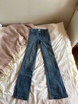 Blå bootcut jeans från Nelly - Snygga blå jeans från Nelly med bootcut passform och två framfickor med lock. Jeansen har dubbla silverknappar i midjan och markerade sömmar längs benen. Perfekt för dig som gillar en retro vibe och vill ha ett par jeans med coola detaljer.