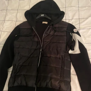 Svart Moncler pufferjacka med huva - Svart pufferjacka från Moncler med stickade ärmar och stor vit zigzagdetalj på ena ärmen. Jackan har huva, dragkedja framtill och två fickor med knappar. Materialmix av polyester och stickat ger en unik look. Perfekt för dig som vill ha något stilrent men ändå sticker ut.