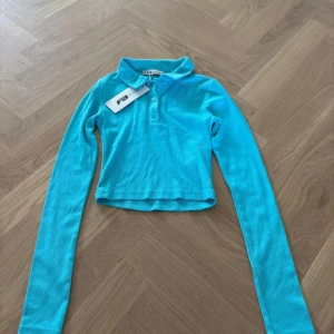 Turkos croppad polotopp från FB Sister - Säljer en turkos croppad polotopp från FB Sister i ribbad bomull. Toppen har lång ärm, klassisk krage och tre knappar framtill. Perfekt för dig som gillar färgstarka och trendiga plagg med snygg passform.