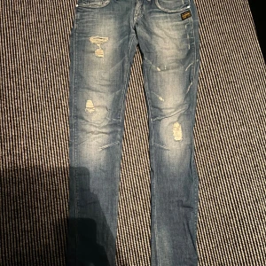 Blå skinny jeans från G-Star RAW - Snygga blå jeans från G-Star RAW med slitna detaljer och skinny passform. Jeansen har klassiska fem fickor, dragkedja och knappstängning samt coola slitningar framtill och bakfickor med zip. Perfekt för dig som gillar streetstyle. De är storlek 27 i midja men passar mig med storlek 29 i midja.